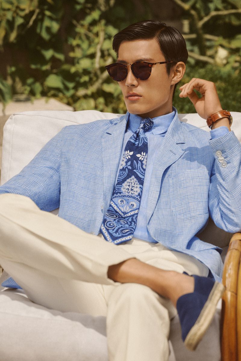 A stylish Asian man in sunglasses, a blue linen blazer, light trousers, and loafers sits outdoors in a relaxed pose.