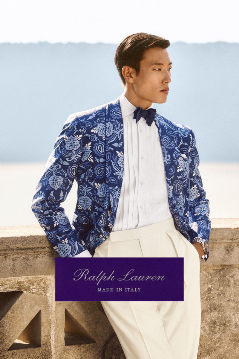 Asian male model poses in a patterned blue blazer and white pants, standing on a stone balcony with a bright sky background.