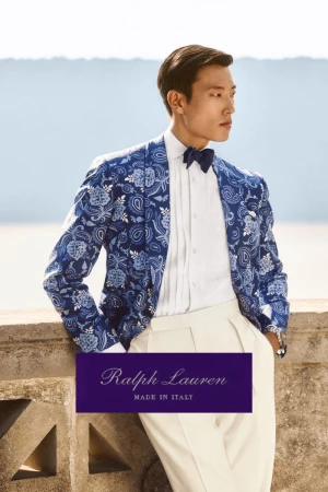 Asian male model poses in a patterned blue blazer and white pants, standing on a stone balcony with a bright sky background.