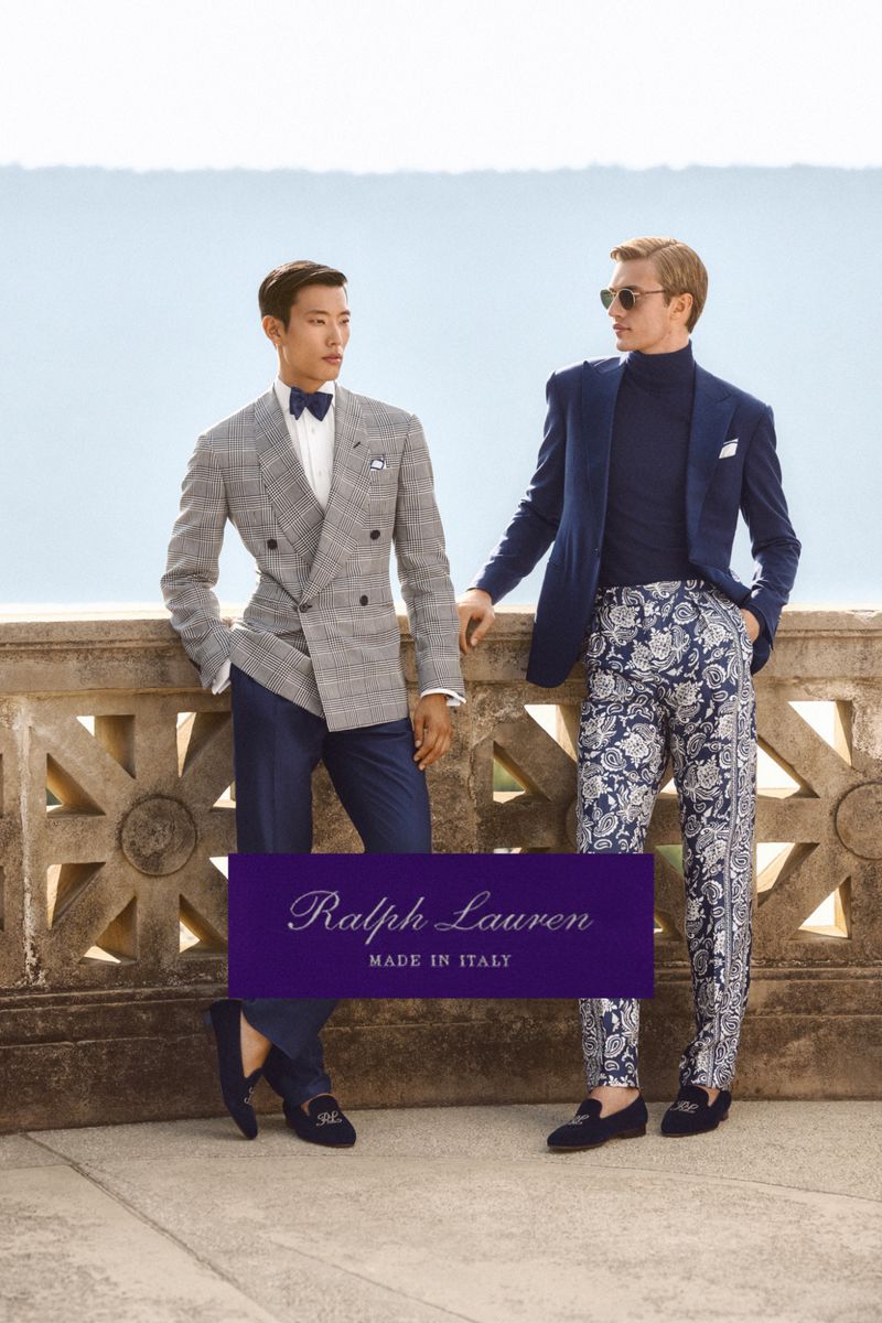 Two men pose outdoors in elegant Ralph Lauren suits and loafers, one in grey check suit with bow tie, the other in floral pants and sunglasses.