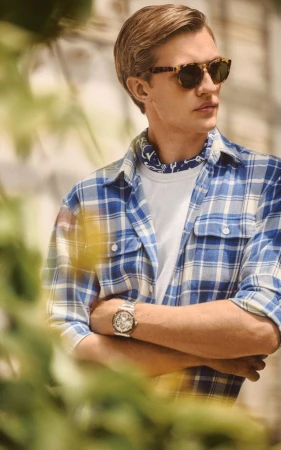 Blond man wearing a blue plaid shirt and sunglasses poses outside with a patterned neckerchief and a wristwatch, looking to the side.