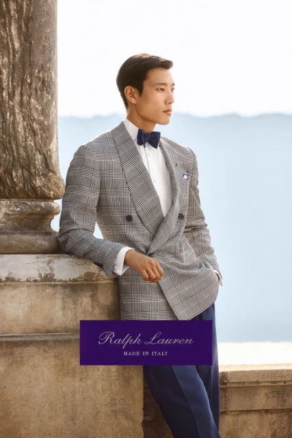 Asian man in grey plaid double-breasted suit with blue bow tie leaning against stone wall with sea view; Ralph Lauren advertisement.