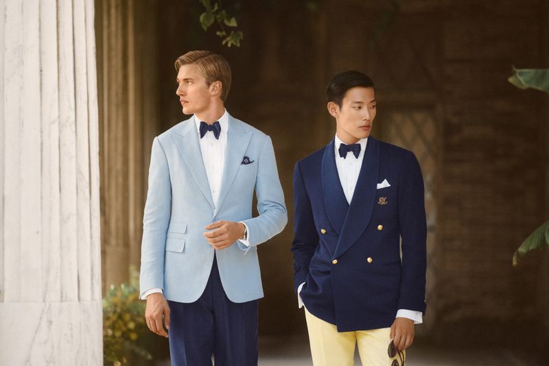Two elegant men in formal wear stand outdoors near columns and greenery, wearing bow ties, jackets, and dress pants for a stylish fashion shot.