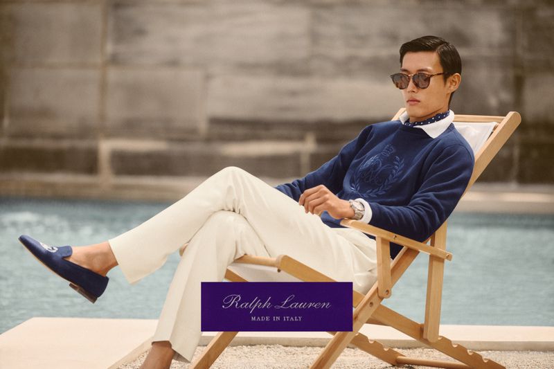 A stylish adult male model lounges in a chair by the water, wearing a navy sweater, white pants, sunglasses, and loafers in a fashion shot.
