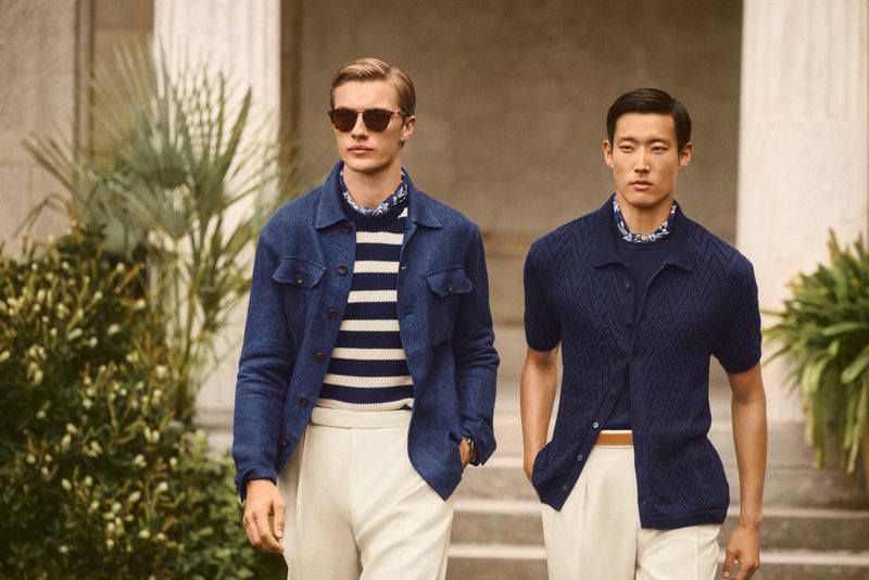 Two male fashion models in navy blue and beige outfits pose outdoors near architecture in a lifestyle fashion shot for social media.