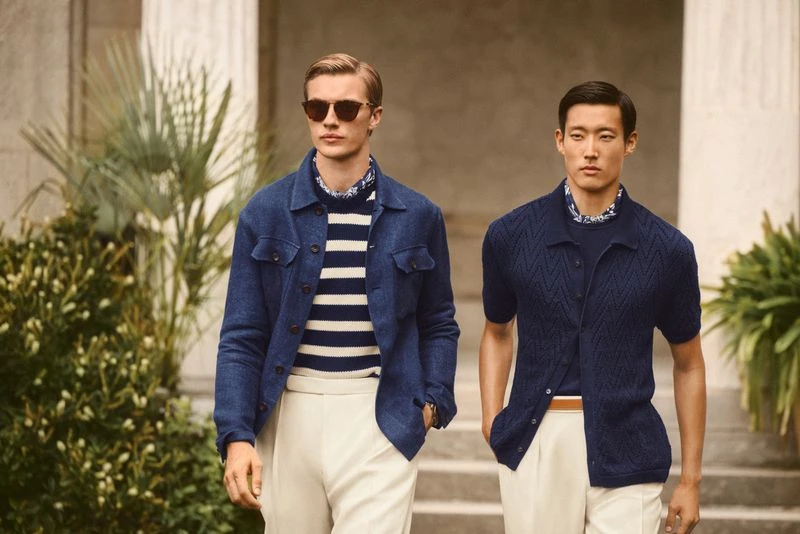 Two male fashion models in navy blue and beige outfits pose outdoors near architecture in a lifestyle fashion shot for social media.