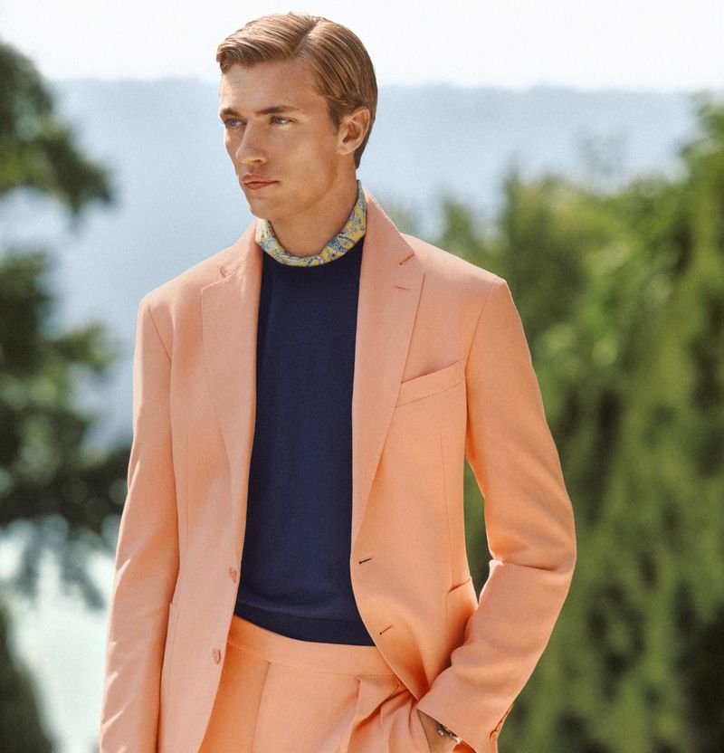 A fair-skinned adult man wears a peach suit with a blue shirt, photographed outdoors with a soft focus background on a sunny day.