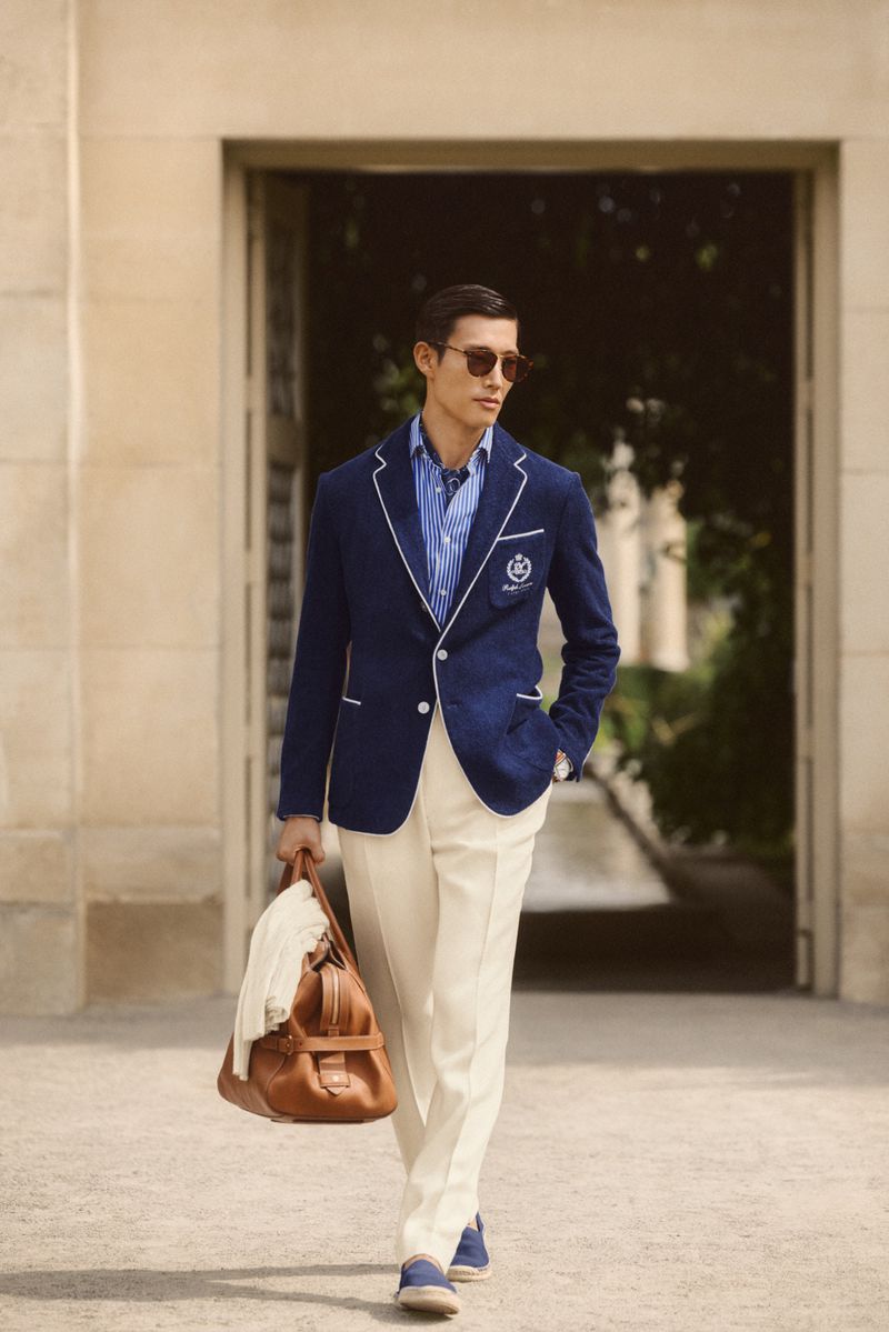 Man in navy blazer and cream-colored pants walks with leather duffel bag, wearing sunglasses on an outdoor paved surface.