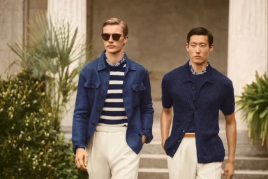 Two male models walking outdoors wearing blue jackets, striped or knit shirts, white trousers, and sunglasses.