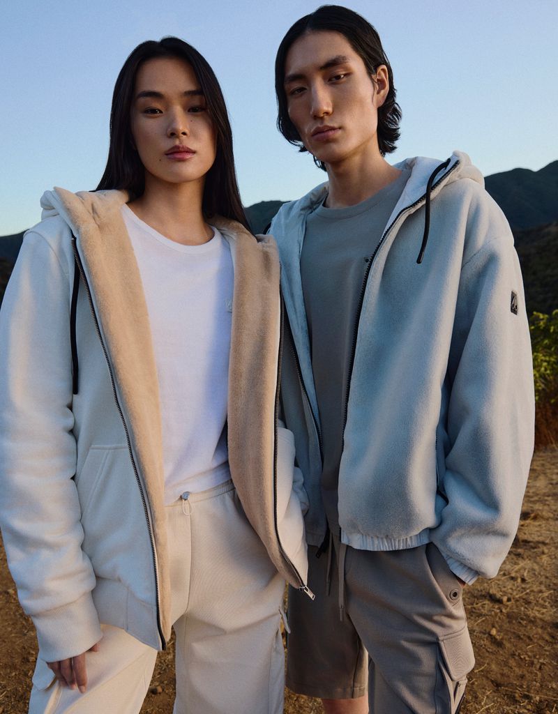 Two models pose outdoors wearing casual hooded jackets and pants, against a natural mountain backdrop under a clear daytime sky.