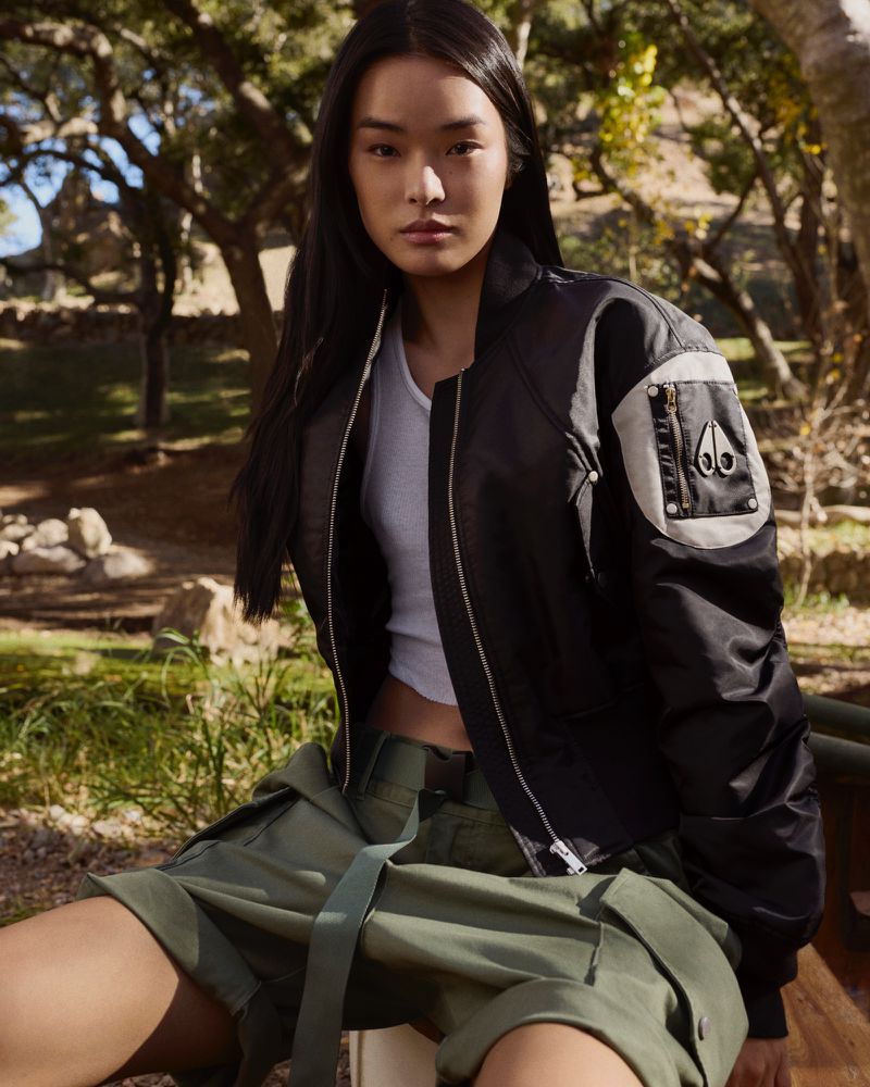 Asian woman in a black bomber jacket and green cargo shorts sits outdoors. The shot captures a relaxed and stylish fashion moment.
