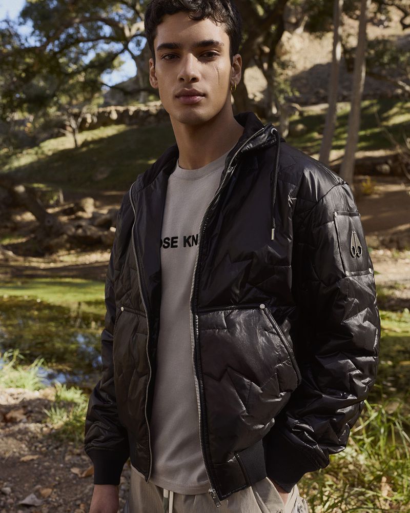 Adult male poses in nature wearing a black quilted jacket with a zipper. T-shirt is visible and the background shows a rural environment.