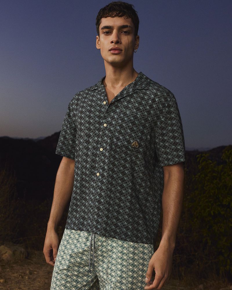Man in patterned short-sleeve shirt and shorts poses outdoors at dusk, showcasing contemporary men's fashion with a natural backdrop.