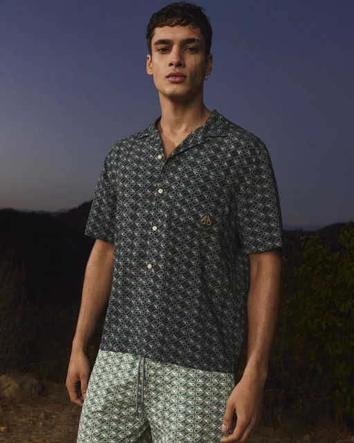 Man in patterned short-sleeve shirt and shorts poses outdoors at dusk, showcasing contemporary men's fashion with a natural backdrop.