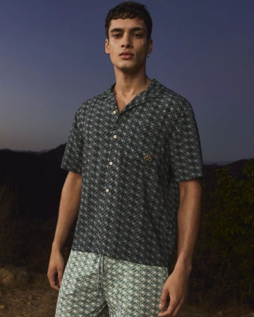 Man in patterned short-sleeve shirt and shorts poses outdoors at dusk, showcasing contemporary men's fashion with a natural backdrop.