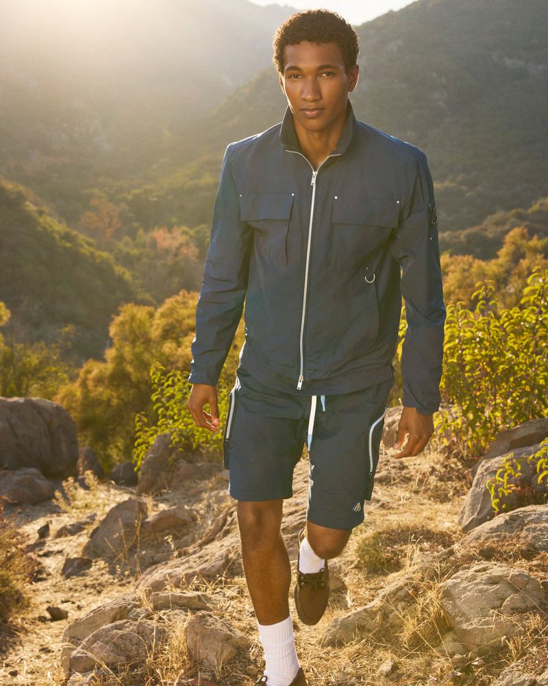 A man in a blue athletic outfit walks on a rocky trail in a sunlit outdoor setting with trees and mountains in the background.