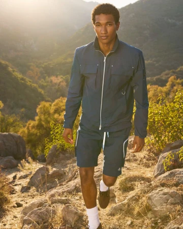 A man in a blue athletic outfit walks on a rocky trail in a sunlit outdoor setting with trees and mountains in the background.