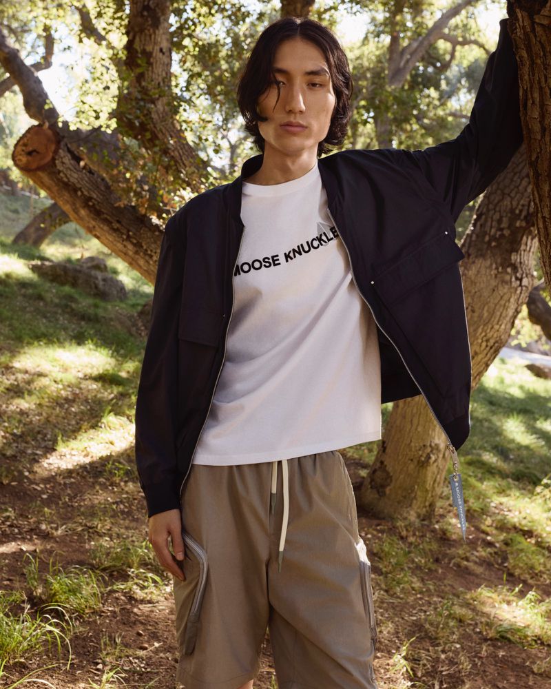 Man poses in a forest wearing a navy bomber jacket, white t-shirt, and khaki shorts. He is leaning against a tree.