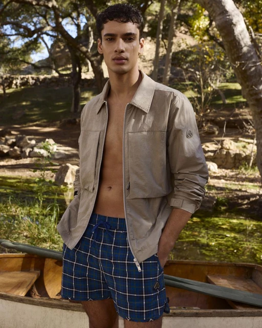 A young adult male stands by a wooden rowboat in a tan jacket and plaid shorts outdoors in natural light, creating a warm, summery feel.