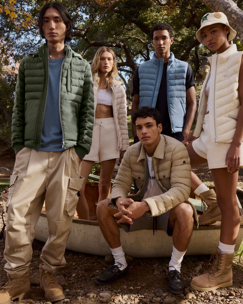 Group of five young adult fashion models in puffer vests and jackets pose together outdoors in a wooded landscape with an old rowboat.