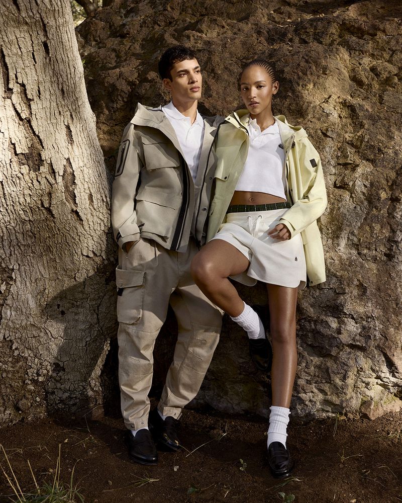 Two fashion models wearing outdoor clothing in a forest setting, showcasing modern apparel and natural light, promoting outdoor lifestyle.