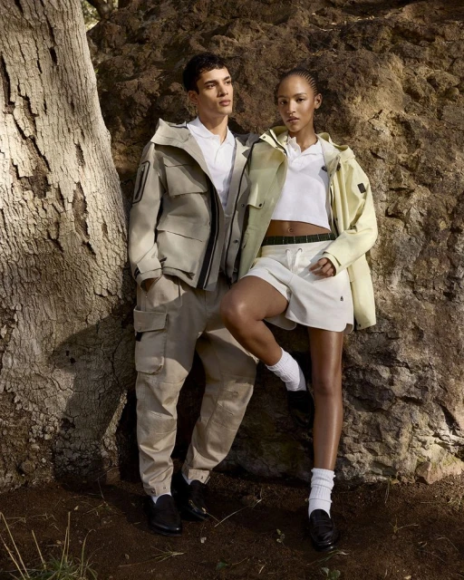 Two fashion models wearing outdoor clothing in a forest setting, showcasing modern apparel and natural light, promoting outdoor lifestyle.