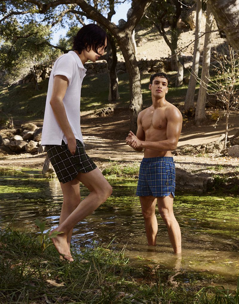 Two men stand in a shallow stream surrounded by trees, one shirtless and one in a polo shirt and shorts, enjoying a sunny day.