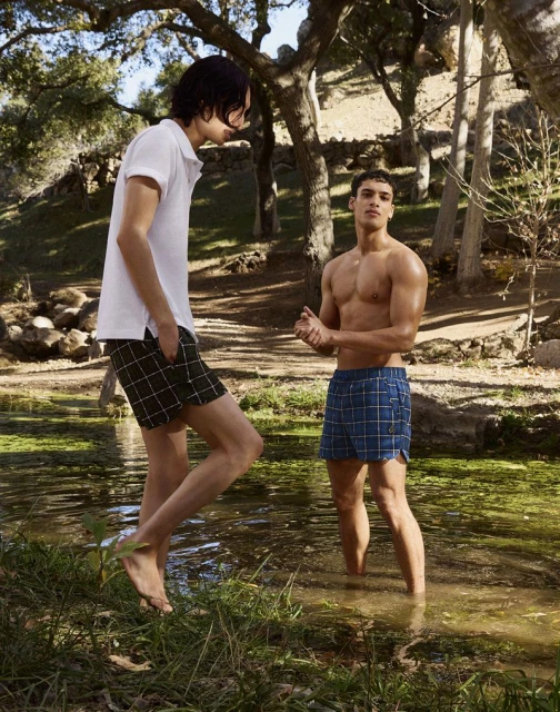 Two men stand in a shallow stream surrounded by trees, one shirtless and one in a polo shirt and shorts, enjoying a sunny day.