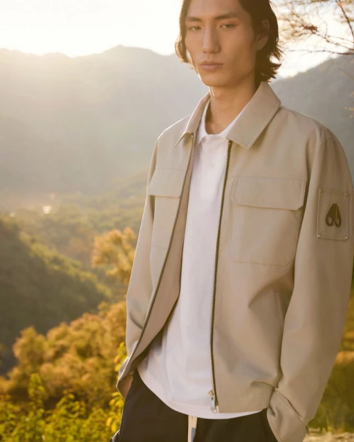 An adult male poses outdoors wearing a beige jacket and white t-shirt. The background features an out-of-focus mountain range.