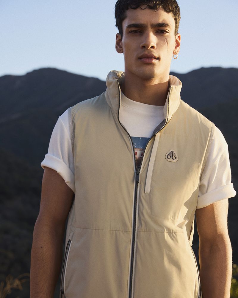 A tan-skinned man in a cream vest stands against a natural mountain backdrop, presenting fashion apparel in natural light.