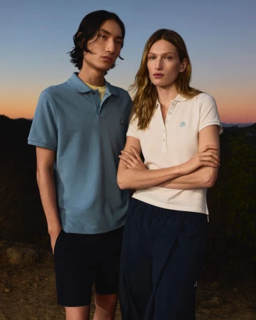 A male and female fashion model stand in an outdoor setting wearing matching polo shirts and shorts at dusk, showcasing lifestyle apparel.