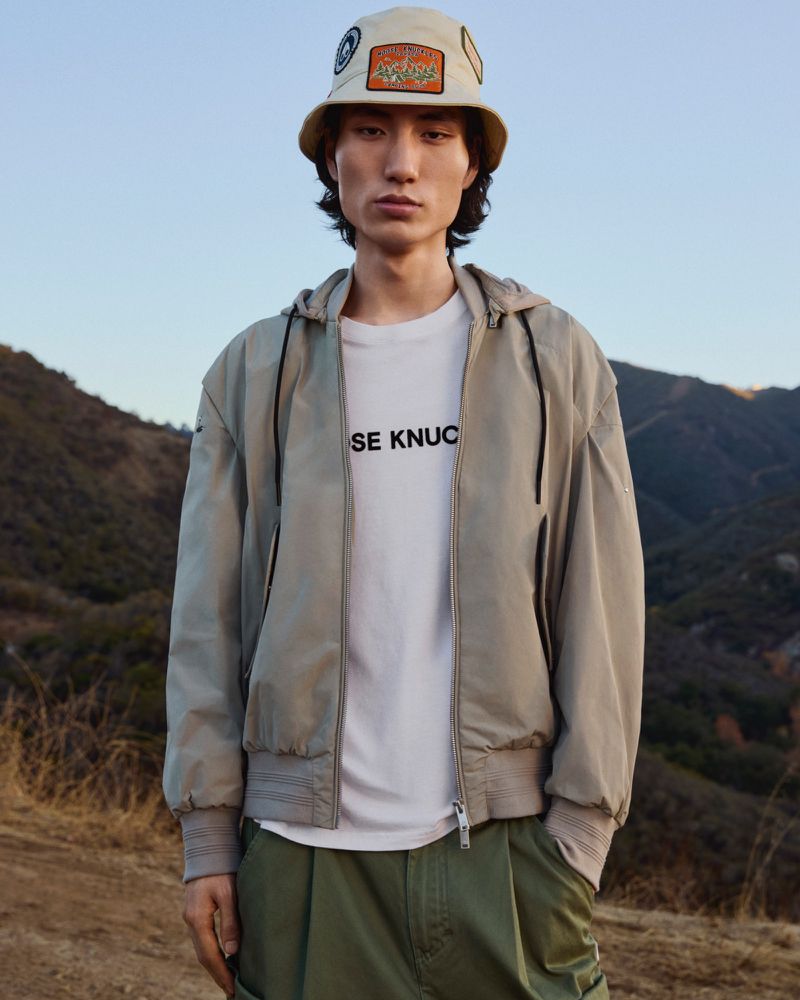 Outdoor fashion shot of a male model in a light grey jacket, white t-shirt, and khaki pants, wearing a bucket hat with patches.