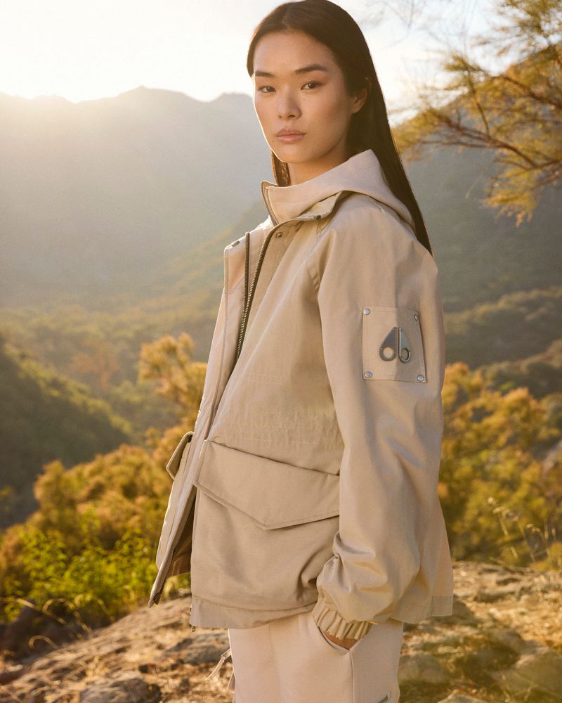 Asian woman in beige hooded jacket stands outside in a nature setting with golden sunlight and background mountain view. Fashion shot.