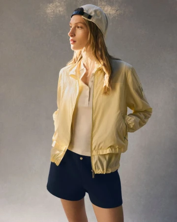 Model wearing a light yellow zip jacket and cap against a textured grey background