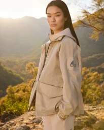 Model wearing a beige hooded jacket with front pockets standing outdoors against a mountainous landscape at sunset.
