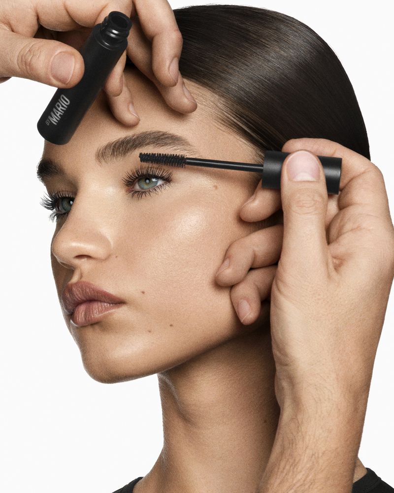 Close-up of an adult female model having mascara applied to her eyelashes by a makeup artist, with natural makeup in a bright setting.