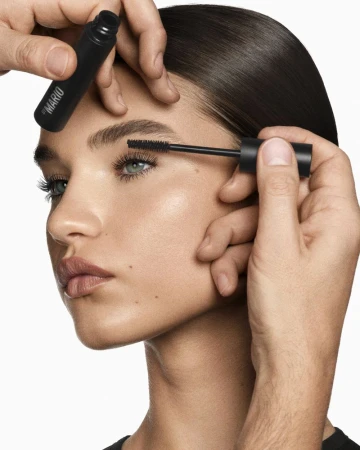 Close-up of an adult female model having mascara applied to her eyelashes by a makeup artist, with natural makeup in a bright setting.