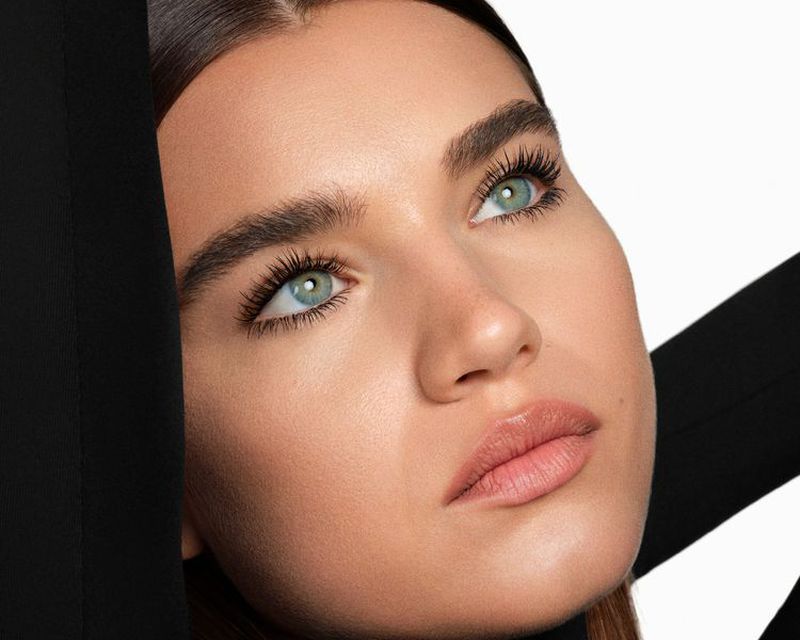 Close-up beauty shot of a light-skinned adult female with light green eyes, wearing a black top against a plain white background.