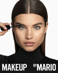 Model applying mascara with a black wand, wearing a black top against a solid white background in the Makeup by Mario campaign.