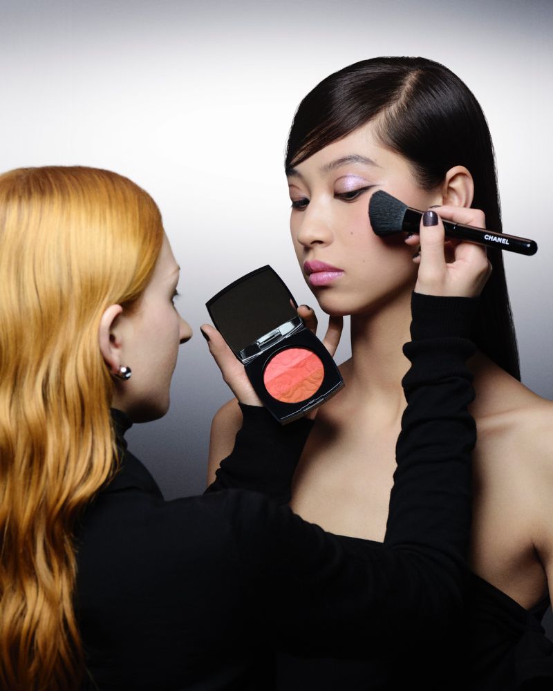 A makeup artist applies blush to a young Asian model in a beauty shot, focusing on cosmetic application and the model's striking features.