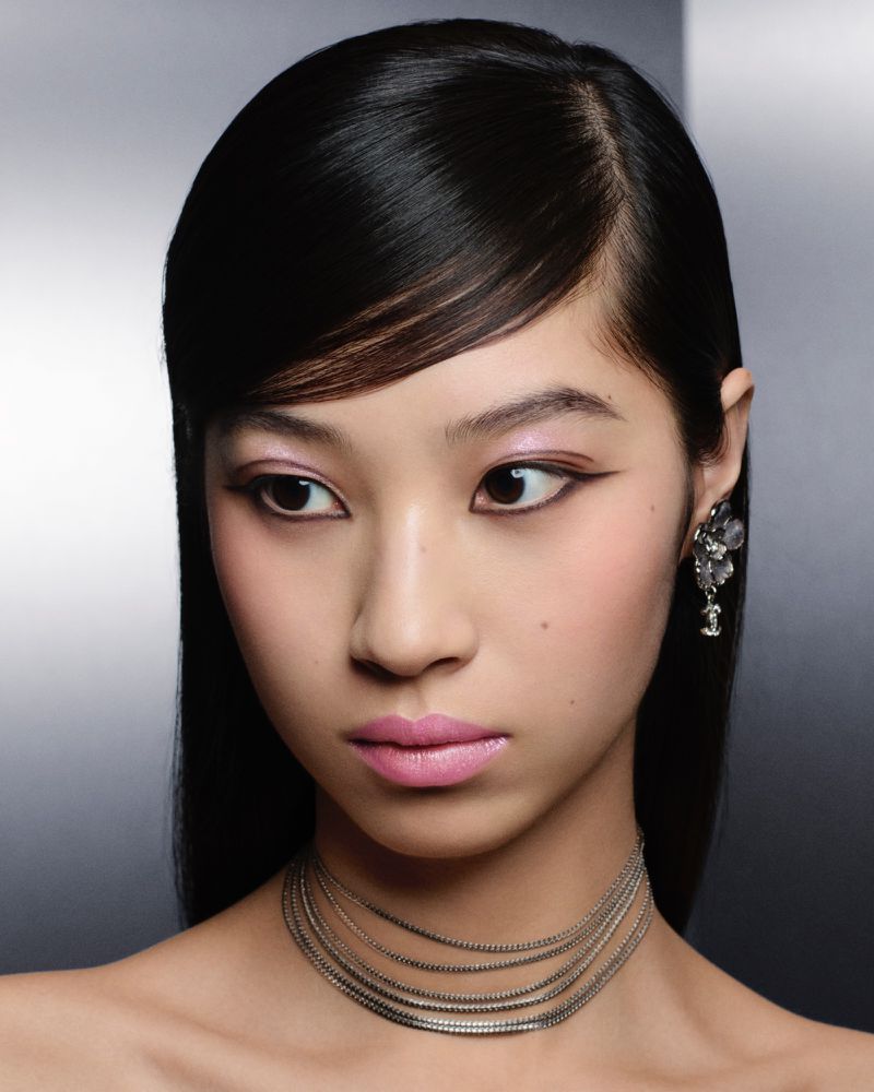 Close-up portrait of a model with sleek dark hair, pink lipstick, and layered silver choker necklace against a smooth grey background