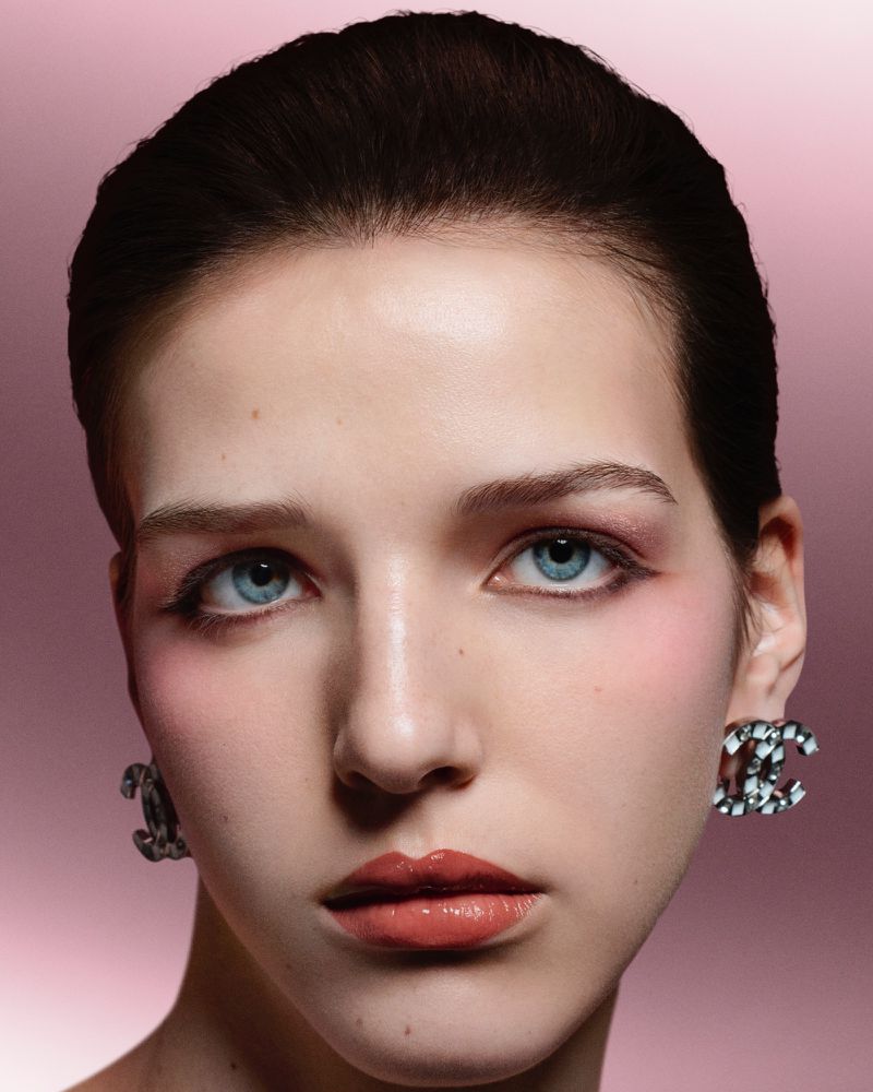 Close-up portrait of a young woman with blue eyes, glossy lips, and statement earrings against a soft pink gradient background.