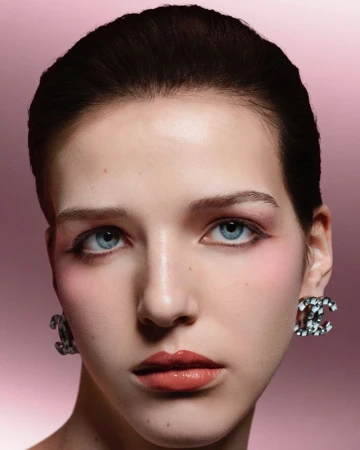 Close-up portrait of a model with blue eyes wearing Chanel CC logo earrings against a pink gradient background.