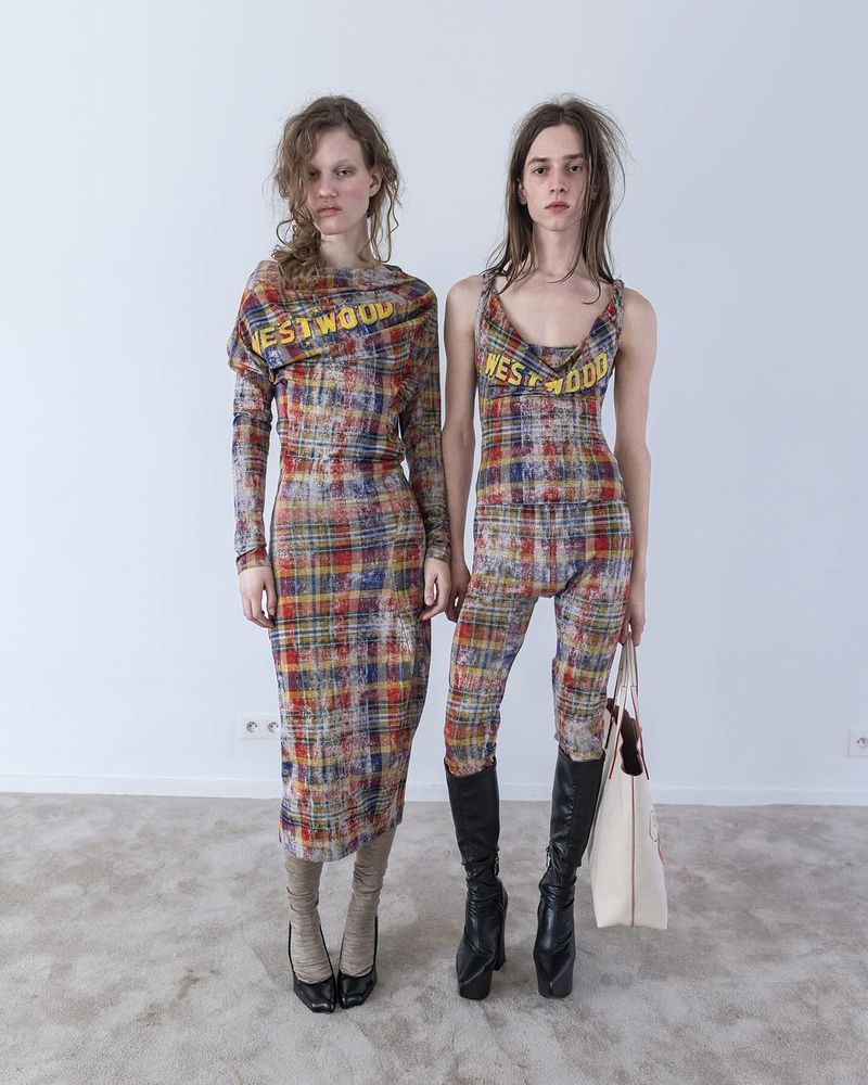 Two fashion models wearing patterned dresses and boots in a bright studio. The dresses have a printed 'Westwood' logo on them.