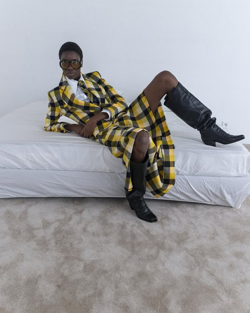 Stylish black woman poses in a yellow plaid suit and black boots, lying on a white bed against a white wall. Fashion shot in a studio.