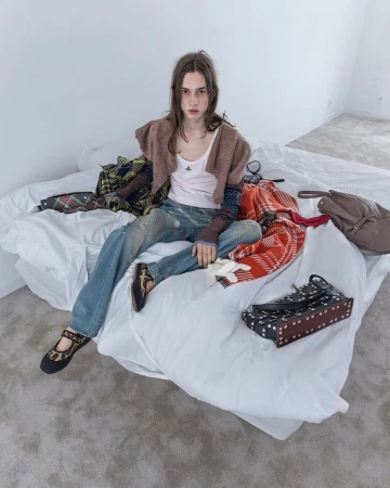 Fashionably dressed young adult sitting on a bed surrounded by clothing and accessories in a minimalist white room, looking at the viewer.