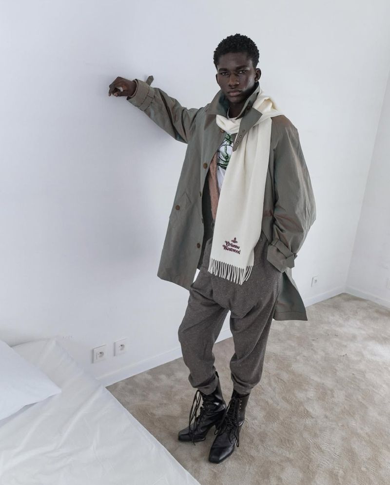 Young man in a light-grey trench coat and cream scarf poses in an indoor setting with a white wall and bed, with a neutral carpeted floor.