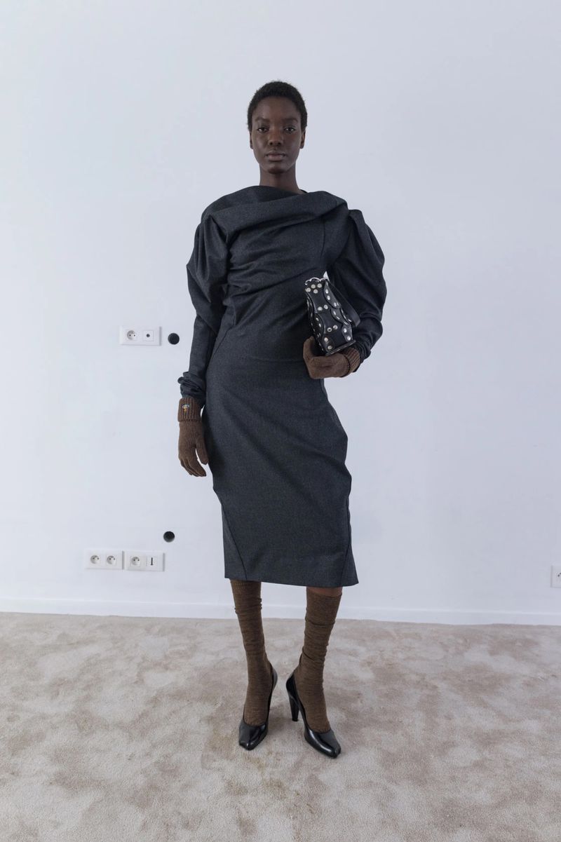 A Black female model poses in a studio, wearing a gray dress with puffy sleeves, brown gloves and hosiery, and black high heels.