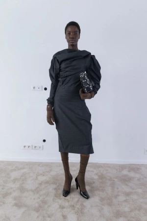 A Black female model poses in a studio, wearing a gray dress with puffy sleeves, brown gloves and hosiery, and black high heels.