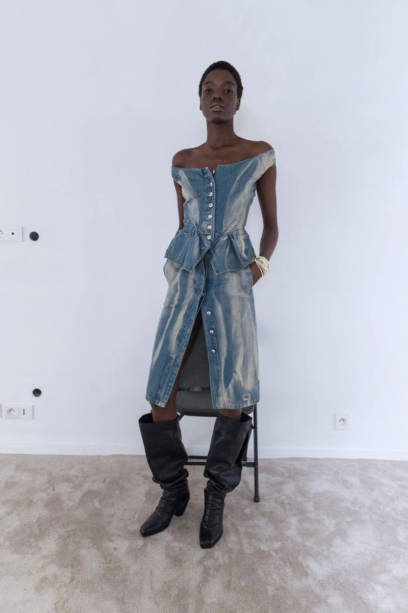 A Black female model poses in a studio, wearing a blue denim dress and black boots, complemented by a pearl bracelet on her left wrist.
